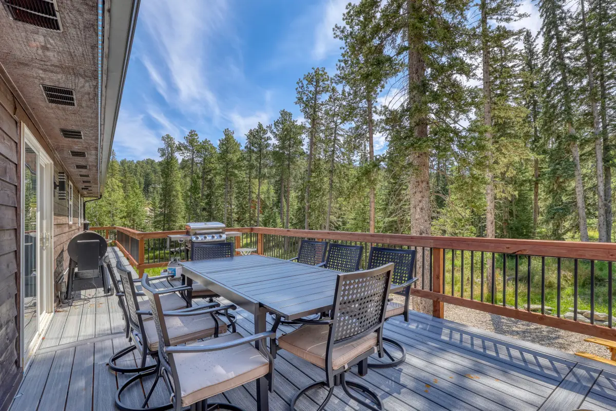 Dining table and chairs on the deck, with a grill at the far end and pines beyond